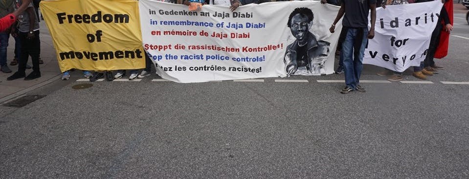 Three front banners hold by people at a protest march. Left one says 'Freedom of Movement', second one 'in Gedenken an Jaja Diabi, stoppt die rassistischen Konktrollen! in remembrance of Jaja Diabi, stop the racist police controls! en mémoire de Jaja Diabi, s'arrête les contrôles racistes', third one 'solidarity for everyone' 
