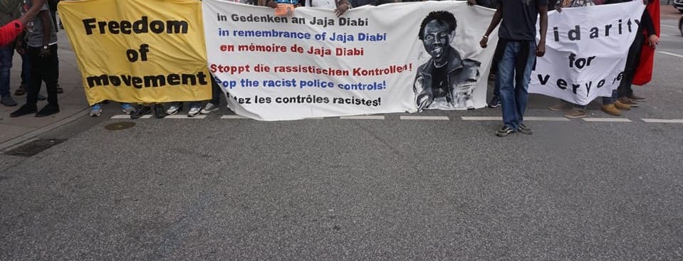Three front banners hold by people at a protest march. Left one says 'Freedom of Movement', second one 'in Gedenken an Jaja Diabi, stoppt die rassistischen Konktrollen! in remembrance of Jaja Diabi, stop the racist police controls! en mémoire de Jaja Diabi, s'arrête les contrôles racistes', third one 'solidarity for everyone'