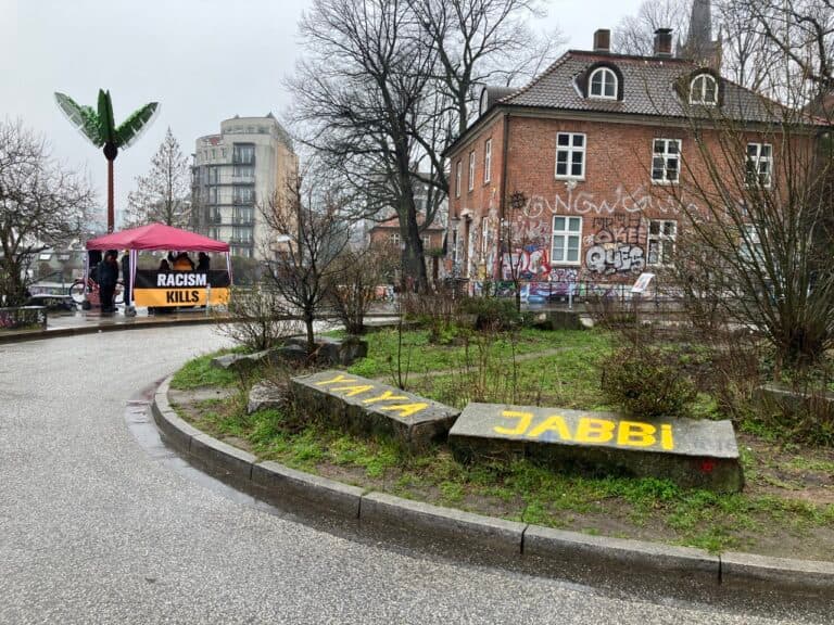 The Yaya Jabbi Circle on February 19, 2023. Two stones in the foreground with Yayas name written on them. Protesters in the background, with a banner 'Racism Kills'